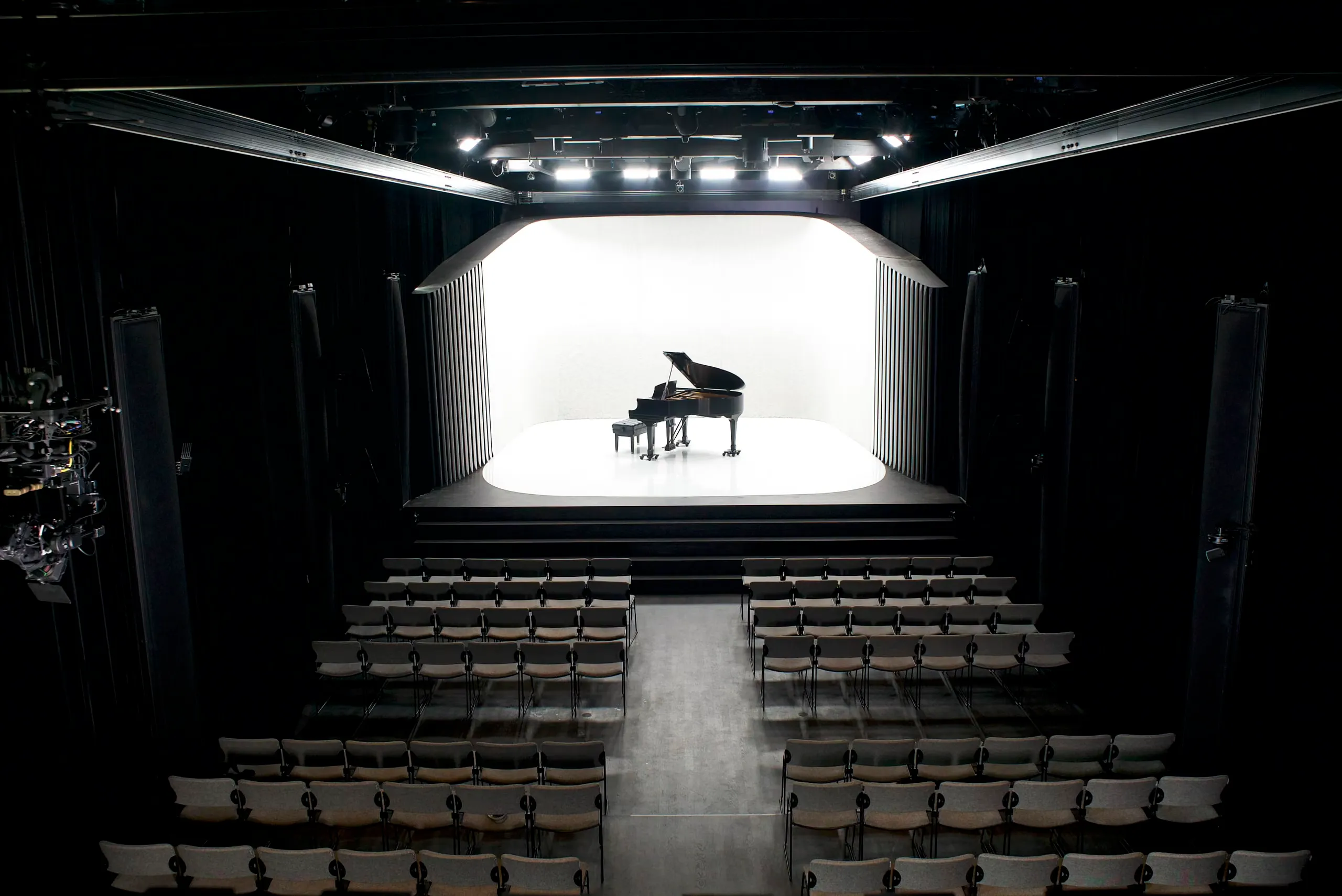 a small concert hall with a grand piano on stage and loudspeaker columns along the walls